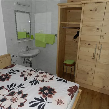 Ein einfaches Zimmer mit einem Bett und einer großen Holzkommode. Im Hintergrund sind ein Waschbecken und Handtücher zu sehen.