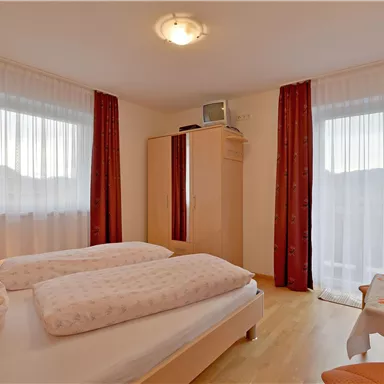 Ein helles Schlafzimmer mit zwei Einzelbetten und großen Fenstern. Die Wände sind in sanften Farben gehalten, und es gibt einen Schrank sowie einen kleinen Tisch mit einer Pflanze.