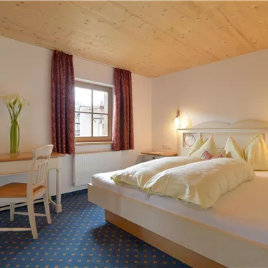 A cozy bedroom with a large bed and bright cushions. The room has a desk and a vase with fresh flowers.