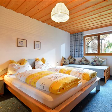 Ein helles Schlafzimmer mit zwei Betten und gelben Bettdecken. Große Fenster lassen viel Licht herein und sorgen für eine gemütliche Atmosphäre.