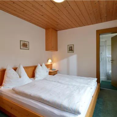 A cozy bedroom with two beds and wooden furniture. The room has a warm atmosphere and a view of an adjoining bathroom.
