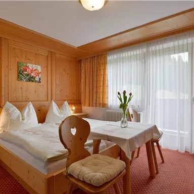 A cozy bedroom with wooden paneling and two beds. A table with fresh flowers is by the window, which lets in plenty of daylight.