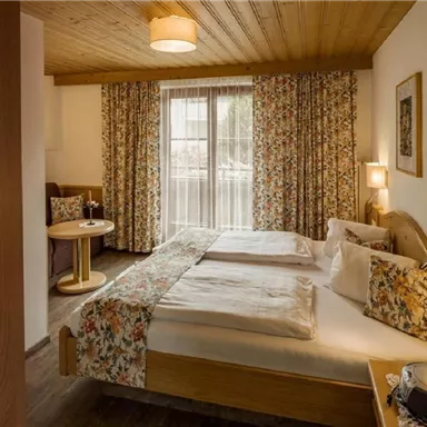 A cozy hotel room with a double bed and floral decor. The room has large windows that let in plenty of light and a comfortable seating area.