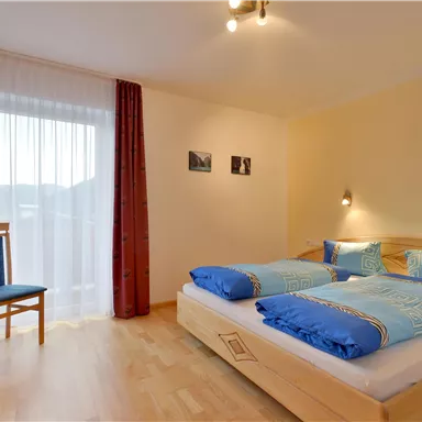 Ein gemütliches Schlafzimmer mit zwei Betten und blauen Bettdecken. Ein Stuhl und ein Fenster mit Vorhängen sorgen für eine helle, einladende Atmosphäre.