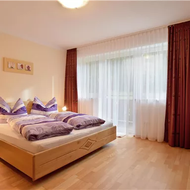 Ein gemütliches Schlafzimmer mit einem Doppelbett und bunten Kissen. Der Raum hat große Fenster mit Vorhängen und einen Fernseher in der Ecke.