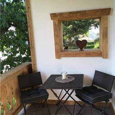 A small balcony with two black chairs and a table. In the background, there is a window overlooking greenery with heart-shaped decor.