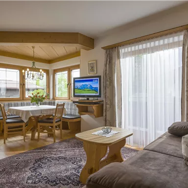 Ein gemütliches Wohnzimmer mit einem Sofa und einem Holztisch. Große Fenster sorgen für viel Tageslicht und Blick auf die Natur.