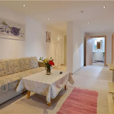 A cozy living room with a beige sofa and a small table. Pictures hang on the wall, and a TV is on a wooden cabinet.