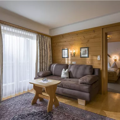 Ein gemütliches Wohnambiente mit einem braunen Sofa und einem kleinen Tisch. Der Raum hat große Fenster mit Vorhängen und einen Zugang zu einem weiteren Zimmer.