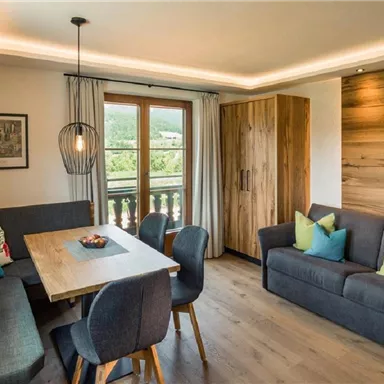 A modern living room with a dining table and comfortable seating. Windows offer a view of the surrounding nature.