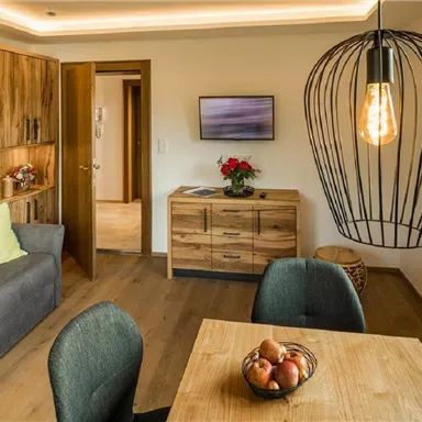 A cozy living space with a gray sofa, a wooden table, and modern chairs. A large lamp hangs over the table, and there are plants and fruit decorations.