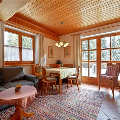 Ein gemütlicher Wohnraum mit Holzmöbeln und einem großen Fenster, das Licht hereinlässt. Der Boden ist mit einem bunten Teppich bedeckt und es gibt einen Tisch mit Stühlen.