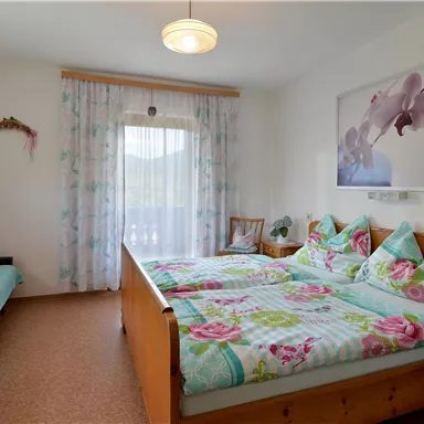 Ein helles Schlafzimmer mit einem großen Doppelbett und bunten Kissen. Es gibt ein Fenster mit Vorhängen und einen gemütlichen Sitzbereich.