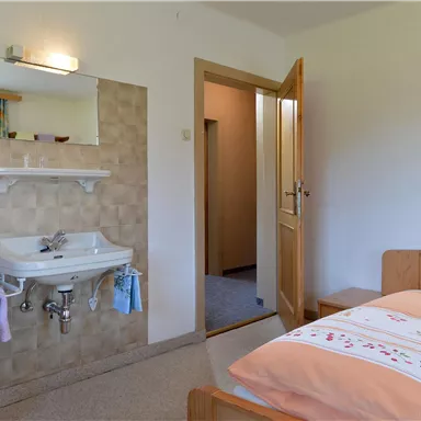 Ein einfaches Zimmer mit einem Bett und einem Waschbecken. Die Tür führt zu einem anderen Raum, und die Wände sind hell gestrichen.