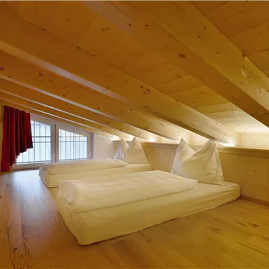 A cozy attic room with wooden ceilings and two beds. Large windows provide plenty of light and a warm atmosphere.