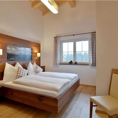 A modern bedroom with a double bed and a wooden panel. The walls are bright, and there is a window with a view as well as a television.
