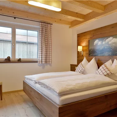 A cozy bedroom with a double bed, wooden decor, and a seating area. Large windows let in plenty of daylight and offer a view outside.
