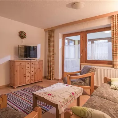 A cozy living room with a couch, a television, and a wooden table. Large windows provide plenty of daylight and a view of the adjoining room.