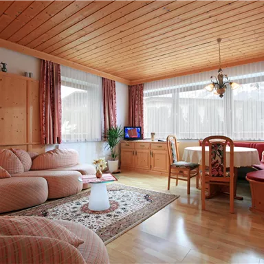 Ein gemütlicher Wohnraum mit Holzdecke und einer großen Fensterfront. Sofas und ein Esstisch bieten Platz zum Entspannen und Essen.