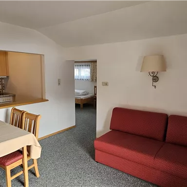 A cozy living room with a red sofa and a dining table. The open kitchen is well-equipped and has wooden cabinets.