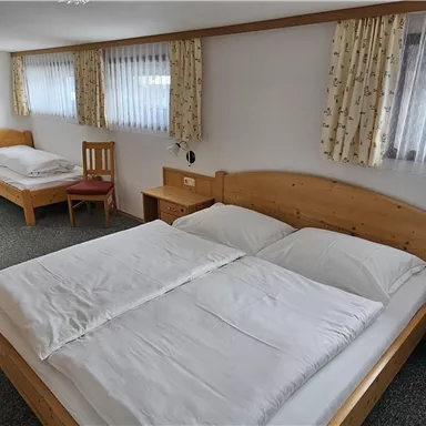 A simple guest room with two beds and a large window. The walls are bright and there is wooden furniture.