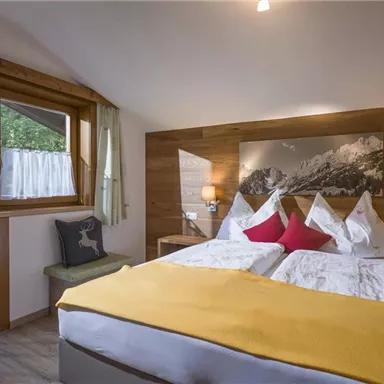 Ein komfortables Schlafzimmer mit einem großen Bett und bunten Kissen. Große Fenster lassen Tageslicht herein und bieten einen Blick auf die Natur.