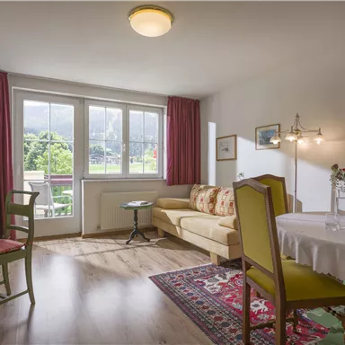 Ein gemütliches Wohnzimmer mit einem Sofa und einem kleinen Tisch. Große Fenster bieten einen Blick ins Grüne.