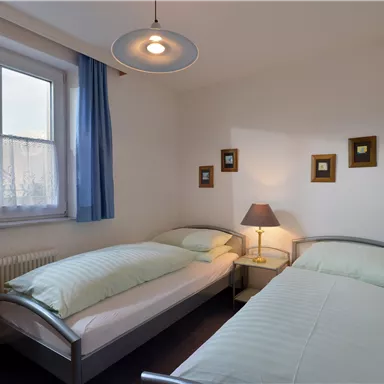 Ein einfaches Schlafzimmer mit zwei Einzelbetten und einem Nachttisch. An der Wand hängen einige kleine Bilder und es gibt ein Fenster mit Vorhängen.