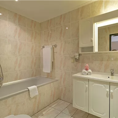 Ein modernes Badezimmer mit einer Badewanne und einem Waschbecken. Die Wände sind hell gefliest und es gibt einen großen Spiegel.