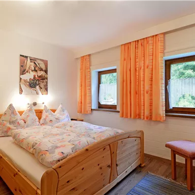 Ein gemütliches Schlafzimmer mit einem Doppelbett und bunten Kissen. Die Fenster sind mit orangefarbenen Vorhängen dekoriert und es gibt eine kleine Sitzecke.