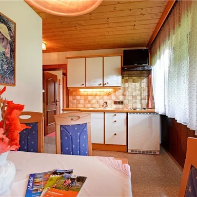 Eine gemütliche Küche mit einem Esstisch, Stühlen und einer Vase mit Blumen. Helle Möbel und ein Fenster mit Vorhängen sorgen für eine einladende Atmosphäre.