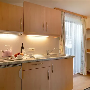 A modern kitchen with wooden cabinets and a sink. A window curtain lets light in, and on the countertop, there is a teapot and fresh fruit.