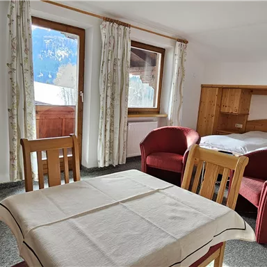 A cozy room with a double bed, two red chairs, and a table. Large windows offer a view of the mountains outside.