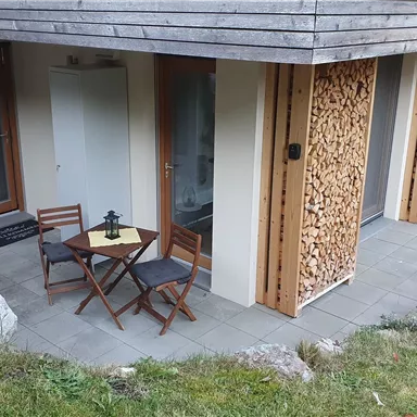 A cozy terrace with two chairs and a small table. The wall is designed with wood paneling, and windows are visible in the background.