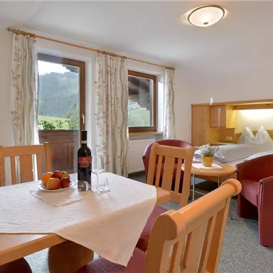 A cozy room with wooden furniture, a table and chairs, and a view of the mountains. On the table, there are fruits and a bottle of wine.
