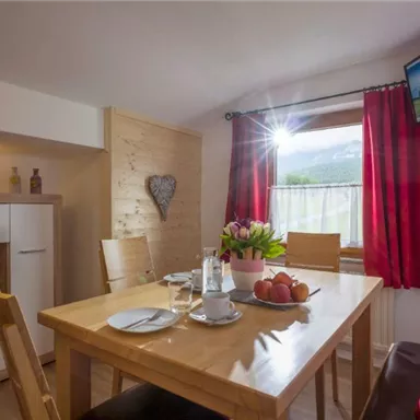 Ein gemütlicher Essbereich mit Holzstühlen und einem Tisch, auf dem frische Blumen und Obst liegen. Licht strömt durch das Fenster und sorgt für eine einladende Atmosphäre.