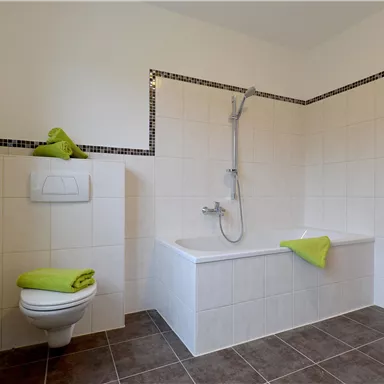 Ein modernes Badezimmer mit einer Badewanne und einer Dusche. Es gibt helle Fliesen und grüne Handtücher auf der Badewanne.