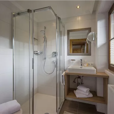 Ein modernes Badezimmer mit einer Glasdusche und einem Holzwaschtisch. Die Fenster lassen viel Tageslicht hereinfallen.