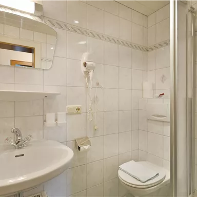 A modern bathroom with white tiles and a sink. There is a shower, a mirror, and a toilet.