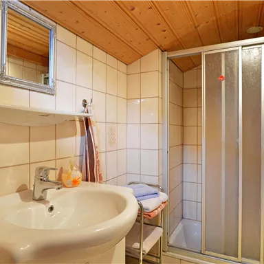 Ein modernes Bad mit einer Dusche und einem Waschbecken. Der Raum hat helle Fliesen und eine Holzdecke.