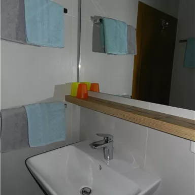 Ein modernes Badezimmer mit einem Waschbecken und Holzregal. Die Handtücher sind ordentlich aufgehängt, und es gibt farbige Becher auf der Ablage.