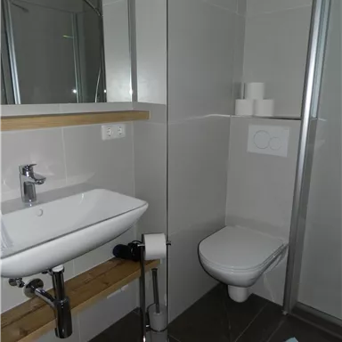 Ein modernes Badezimmer mit Waschtisch, Toilette und Dusche. Die Wände sind hell gefliest und die Einrichtung ist schlicht und funktional.