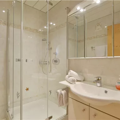 Ein modernes Badezimmer mit einer Glasdusche und einem Waschbecken. Die Wände sind hell gefliest, und es liegen frische Handtücher bereit.