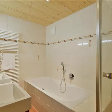 A modern bathroom with a bathtub and a separate shower. The walls are tiled in light colors, and there are two sinks.