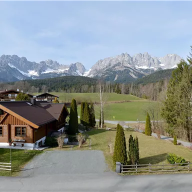 Eine idyllische Landschaft mit einem Holzhaus im Vordergrund und majestätischen Bergen im Hintergrund. Die Szene zeigt grünes Land und Bäume unter einem klaren Himmel.