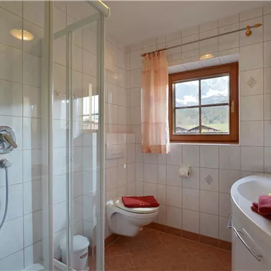 Ein modernes Badezimmer mit einer Dusche, einem WC und einem Waschtisch. Große Fenster lassen viel Licht herein und die Handtücher sind in Rot gehalten.