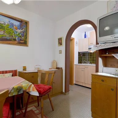 A cozy dining area with a table, chairs, and a picturesque mural. In the background, a small kitchen and a television are visible.