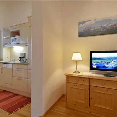 Eine moderne Küche mit Holzschränken und einem kleinen Essbereich. Im Raum befindet sich ein Fernseher auf einer Anrichte und ein Fenster, das Tageslicht hereinlässt.