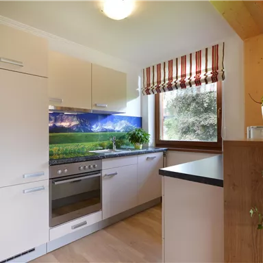 Eine moderne Küche mit hellen Schränken und einem großzügigen Fenster. Im Hintergrund ist eine Landschaft mit Bergen zu sehen.