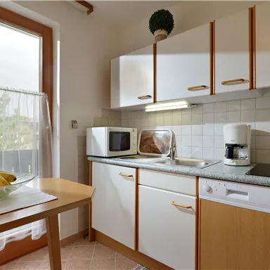 Eine moderne Küche mit weißen Schränken und einer großen Fensterfront. Ein Tisch mit einer frischen Obstschale ist im Vordergrund zu sehen.